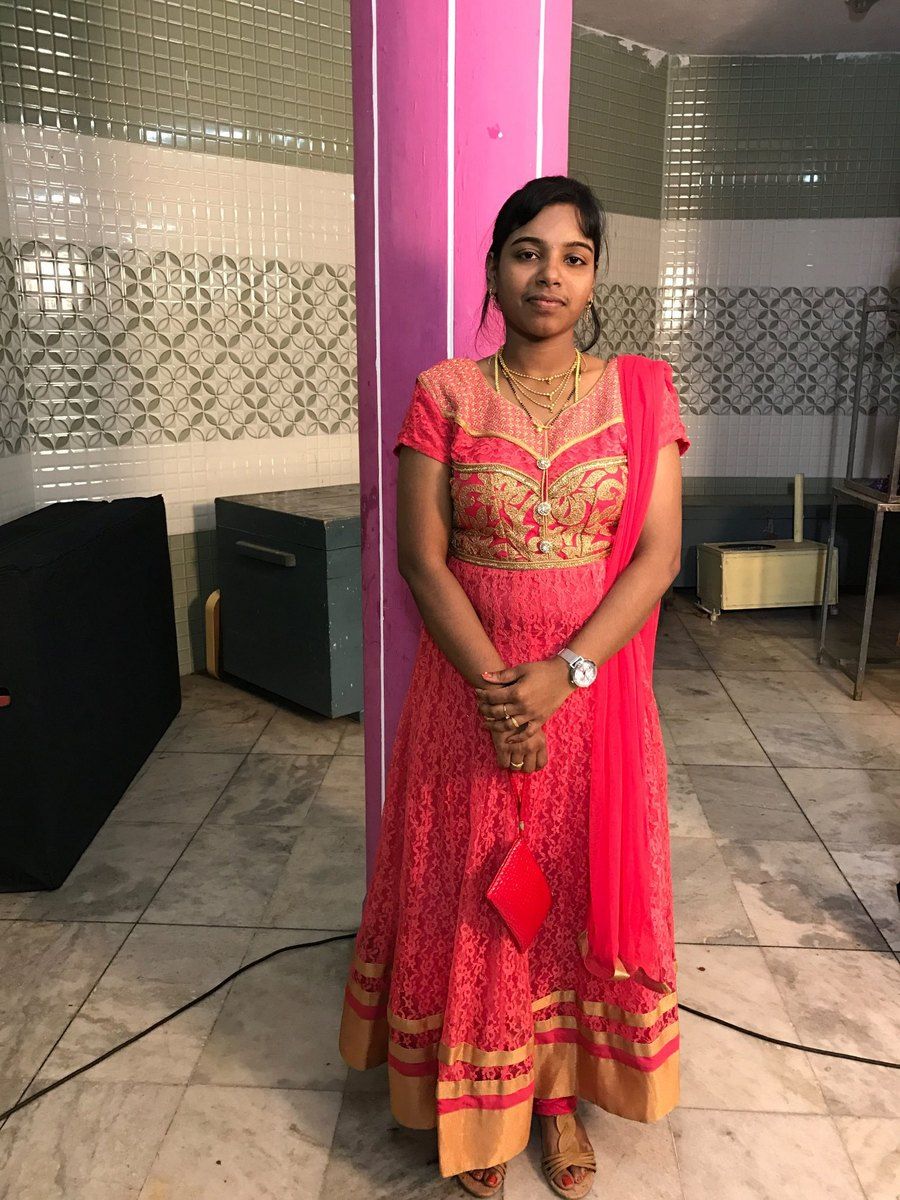 Nithya partway through her transformation, in the same coral-pink dress, standing by a pink pillar
